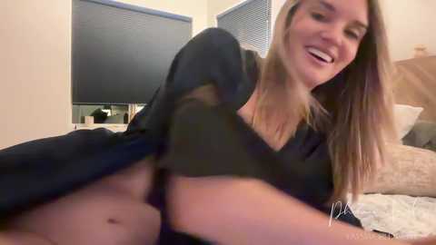 Video of a smiling Caucasian woman with long, blonde hair, wearing a black shirt, lying on a bed in a cozy bedroom with beige walls and a window with blinds.