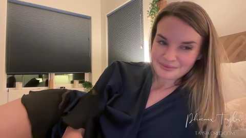Video of a smiling Caucasian woman with light skin, straight brown hair, and a slender build, wearing a dark navy top, sitting on a bed with a white duvet. The background features a window with closed blinds and a potted plant.