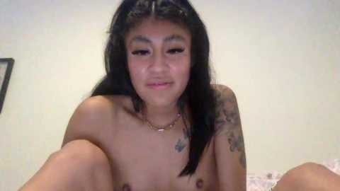 A video of a topless young Asian woman with long black hair, tattoos, and a small necklace, sitting on a bed with white sheets, smiling.
