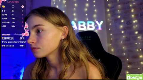 Video of a young Caucasian woman with long brown hair, wearing a strapless top, sitting in a black gaming chair. Background includes a brick wall with fairy lights and the word \"ABBY\" in blue and white letters. Streaming platform interface with live chat on the left.