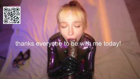 Video of a young blonde woman in a shiny, black latex suit, kneeling on a bed, giving a blowjob to an erect, black dildo, with text overlay thanking viewers.