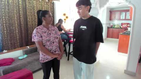 A video shows a middle-aged woman with dark hair and a pink floral blouse, standing with a teenage boy in a living room. The background features a dark curtain, a sofa, and a kitchen with red cabinets.