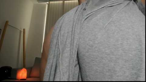 Video of a person's muscular back in a gray T-shirt, standing in a dimly lit room with beige curtains, a wooden chair, and a red lantern on the floor.