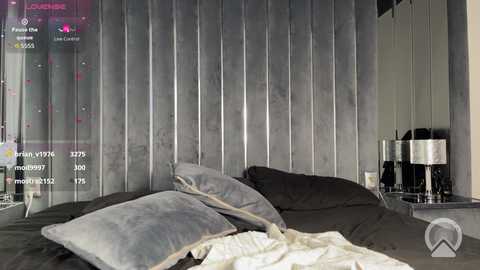 A video of a modern, minimalist bedroom with a plush, gray velvet headboard, a bed adorned with gray and white pillows and sheets, and a silver metallic bedside table with a lamp.
