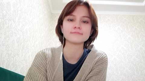 Video of a young woman with light skin, short brown hair, wearing a beige knitted cardigan over a dark blue shirt, standing against a textured white wallpaper background.