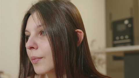 Media: A close-up video of a young woman with straight, dark brown hair and fair skin, smiling gently. She is indoors, with a blurred kitchen background featuring appliances.