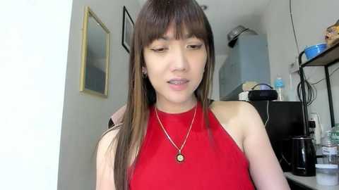 A video of a young Asian woman with long, straight brown hair and a slim build, wearing a red sleeveless top and a necklace, standing indoors with a plain background.