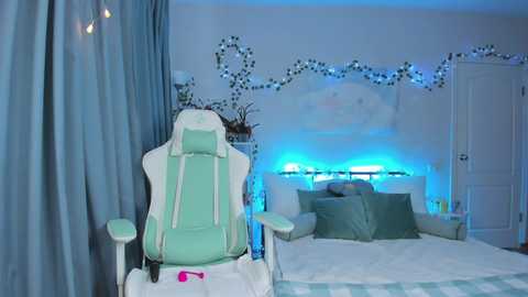 Video of a cozy, blue-themed bedroom with a light green gaming chair, white bed, green pillows, fairy lights, and a white door.
