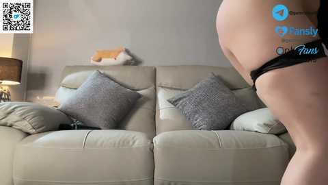Video of a fair-skinned woman in black lingerie bending over a beige leather couch, with two gray throw pillows and a beige blanket, in a dimly lit living room.