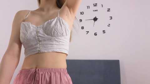 Media: Video of a young woman with light skin, wearing a light gray, lacy bralette and pink shorts, raising her arm, standing in a minimalist room with a white wall and a blue clock.