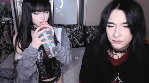 Video of two young women with pale skin, long black hair, and gothic fashion, drinking from colorful plastic bottles in a dimly lit room.