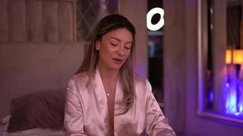 Media: Video of a blonde woman in a satin robe, sitting on a bed with a window and a lit ring light in the background, dim lighting.