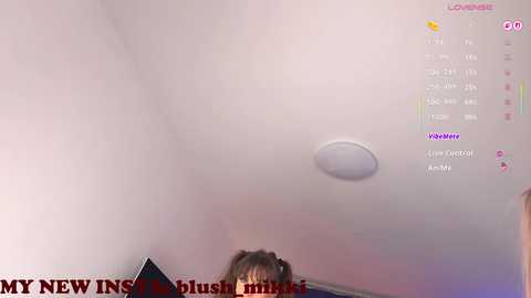 A video captures a young woman with brown hair, partially visible, in a dimly lit room with a white ceiling and a small, round ceiling light.