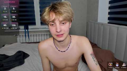 Video of a young, slender, shirtless boy with messy blonde hair, wearing a beaded necklace, sitting on a bed with white sheets in a dimly lit room.