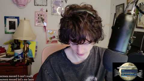 Video of a young person with curly brown hair, wearing a dark gray T-shirt, sitting in a chair in a cluttered room with framed photos and a lamp on a wooden desk.