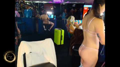 Video of a dimly lit airport lounge with people in casual attire, a white jacket draped over a chair, and a green suitcase.