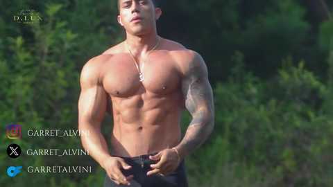 Video of a muscular, shirtless man with tattoos, wearing black pants, standing outdoors in a forest. He has a confident expression. The background shows lush green foliage.