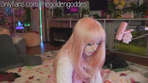 Media: Video of a young woman with long, straight, pink hair, wearing a pink outfit, sitting on a colorful blanket, in a cozy, dimly-lit room with a gaming setup in the background.