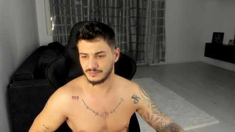 Video of a shirtless, muscular man with short dark hair and tattoos, sitting on a black chair in a modern, minimalist living room with gray walls, a black couch, and a white rug.