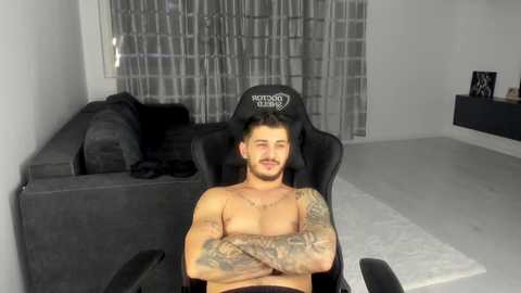 Video of a muscular, shirtless man with tattoos sitting in a black gaming chair, arms crossed, in a minimalist living room with gray curtains and a dark gray sofa.