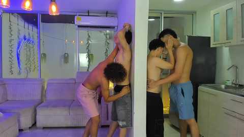 Video of three shirtless young men, two embracing, in a modern apartment with white walls, a sofa, and kitchen.