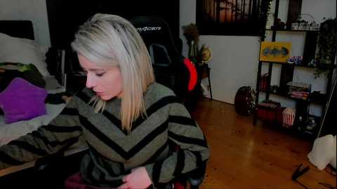 Video of a young blonde woman in a striped sweater, seated at a wooden desk in a cluttered bedroom with a gaming chair, guitar, and books.