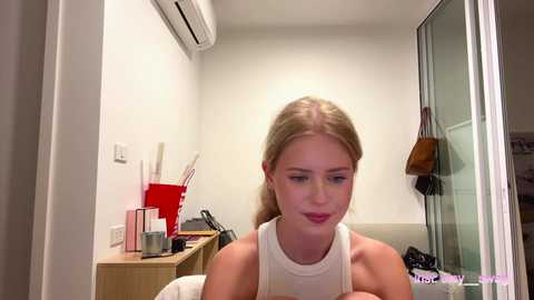 Video of a blonde woman in a sleeveless white top, sitting in a small, modern room with minimalistic decor. A red container and other items are on a wooden shelf behind her.