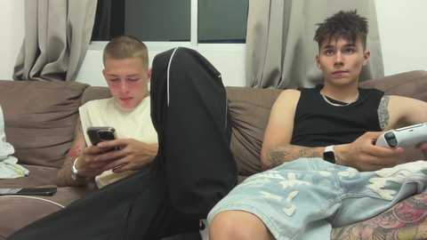 Media: Video of two young men, one with a shaved head, sitting on a brown couch, playing video games, wearing casual attire, with a black duffel bag between them.