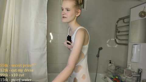 Video of a slender, fair-skinned woman with short blonde hair, wearing a wet, white, polka-dotted dress, standing in a modern bathroom with a shower, towel rack, and toiletries.