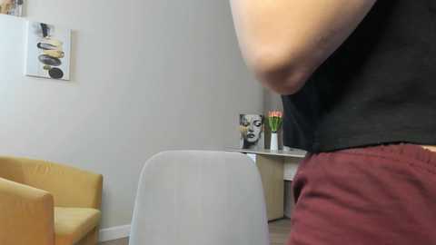 Video of a person in a black tank top, maroon shorts, standing beside a white chair in a minimalist room with beige walls, yellow chair, and abstract art.