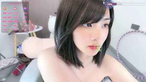 A high-resolution, 3D-rendered photo of an Asian woman with straight black hair and fair skin, wearing a strapless top, lying on a bed with a white sheet. She has a gag in her mouth, and the room is cluttered with toys and a pink hoop.