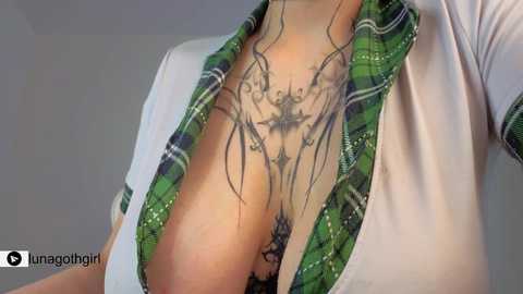 A close-up video of a woman's chest, showing a detailed, intricate tattoo of a spider web covering her right breast, partially visible through an unbuttoned green plaid shirt.