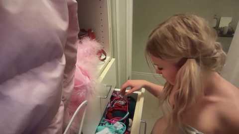 Video of a blonde-haired girl in a strapless top, reaching into a pink, fluffy, open closet, sorting through colorful clothing.