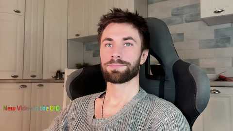 Video of a young, bearded man with short dark hair in a gaming chair, wearing a gray sweater. Background shows a modern kitchen with light cabinets and a tiled backsplash.