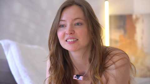 Video of a young Caucasian woman with long, straight brown hair, wearing a pink necklace, smiling. She's in a softly lit bedroom with a white bed and blurred, abstract wall art in the background.