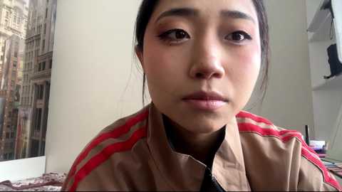 Video of an Asian woman with fair skin, dark hair tied back, wearing a beige jacket with red stripes, looking serious in a modern, bright room with a cityscape mural.