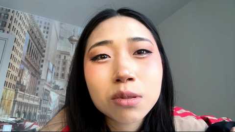 Video of an Asian woman with long black hair, light skin, and subtle makeup, pouting slightly, against a mural backdrop of New York City.