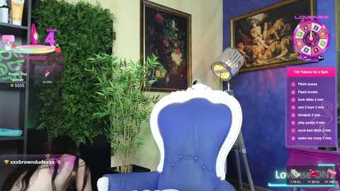 Media: Video of a modern, vibrant room with a blue, tufted armchair, green potted plants, and classical paintings on beige walls. A \"Love Island\" game screen with contestants' faces is displayed in the background.