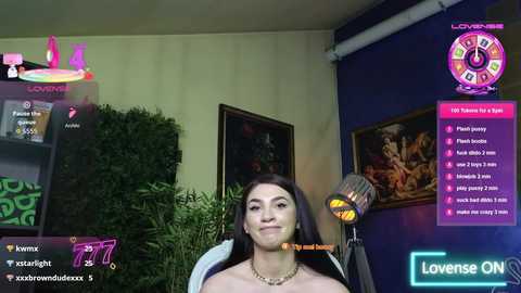 Media: Video of a livestream featuring a woman with long dark hair, wearing a necklace, sitting in a cozy room with greenery, blue walls, and a TV displaying game information.