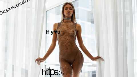 Video of a slender, Caucasian woman with long blonde hair, wearing a fishnet bodysuit, standing in a brightly lit, modern room with white curtains.