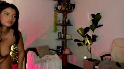 Video of a topless Latina woman with long dark hair, standing in a dimly lit, cluttered room with a pink and red glow, featuring a green plant, shelves, and a white chair.