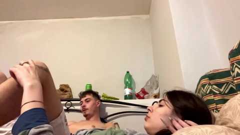 Video of a man and woman in bed; man shirtless, woman in glasses, both facing each other, bed messy with green bottle and stuffed animal.