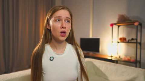A video of a young, fair-skinned woman with long, straight brown hair, wearing a white t-shirt and looking surprised. She's in a modern bedroom with beige curtains, a desk with a lamp, and a shelf with books.