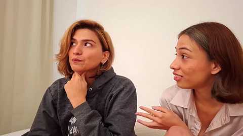 Video of two women in casual attire; one with short brown hair, the other with long brown hair, speaking indoors with beige walls and curtains.