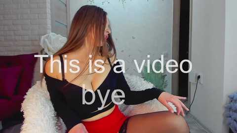 Video of a brunette woman with a curvy figure in a black off-shoulder top and red shorts, sitting on a white fur rug in a modern living room with brick walls. Text overlays read \"This is a video by bye.\