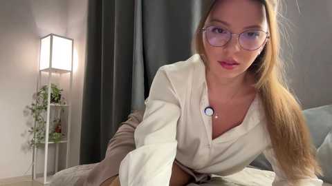 Media: Video of a Caucasian woman with long blonde hair, wearing glasses, a white blouse, and a stethoscope, sitting on a bed in a modern, softly lit room with a grey curtain.