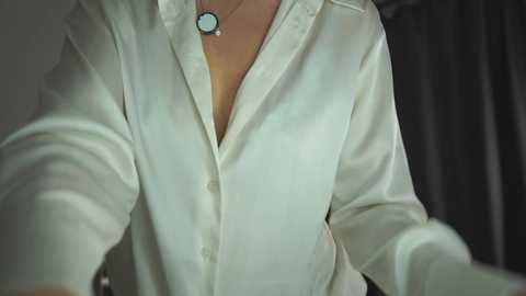 Media: Video of a person wearing a white button-up shirt, featuring a turquoise pendant necklace. The background is blurred, with dark, possibly velvet curtains. The shirt is slightly unbuttoned, revealing a hint of cleavage.