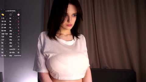 Video of a young woman with shoulder-length black hair, wearing a white cropped T-shirt, standing in front of a dimly lit curtain backdrop.