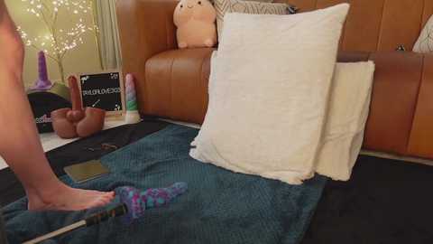 A video shows a person's legs and feet wearing colorful, patterned socks on a teal rug in a cozy, dimly-lit living room with a brown leather couch, plush toys, and fairy lights.