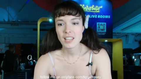 Video of a young woman with straight brown hair in pigtails, wearing a pink spaghetti-strap top, smiling. Background shows a dimly-lit, cluttered room with a yellow object and a blue sign.